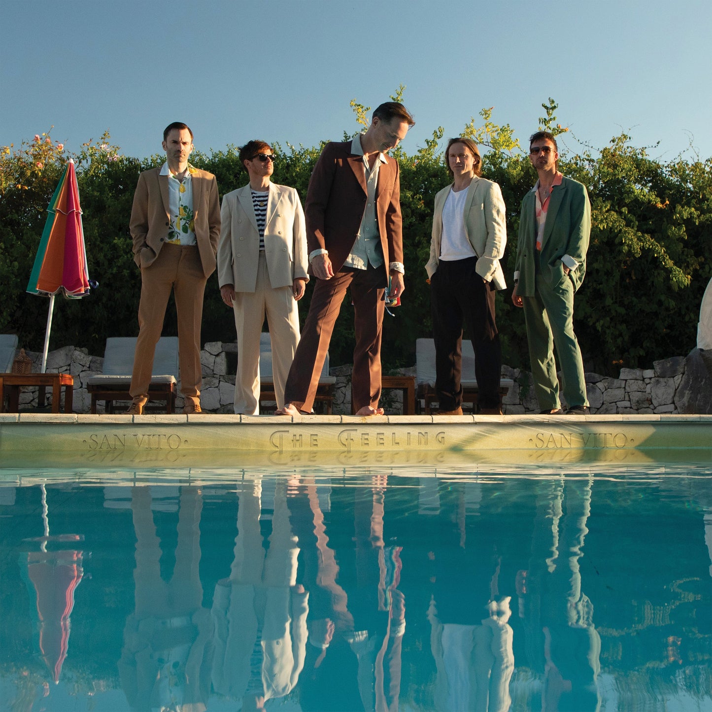 Five men in suits standing by a swimming pool, reflected in the water.