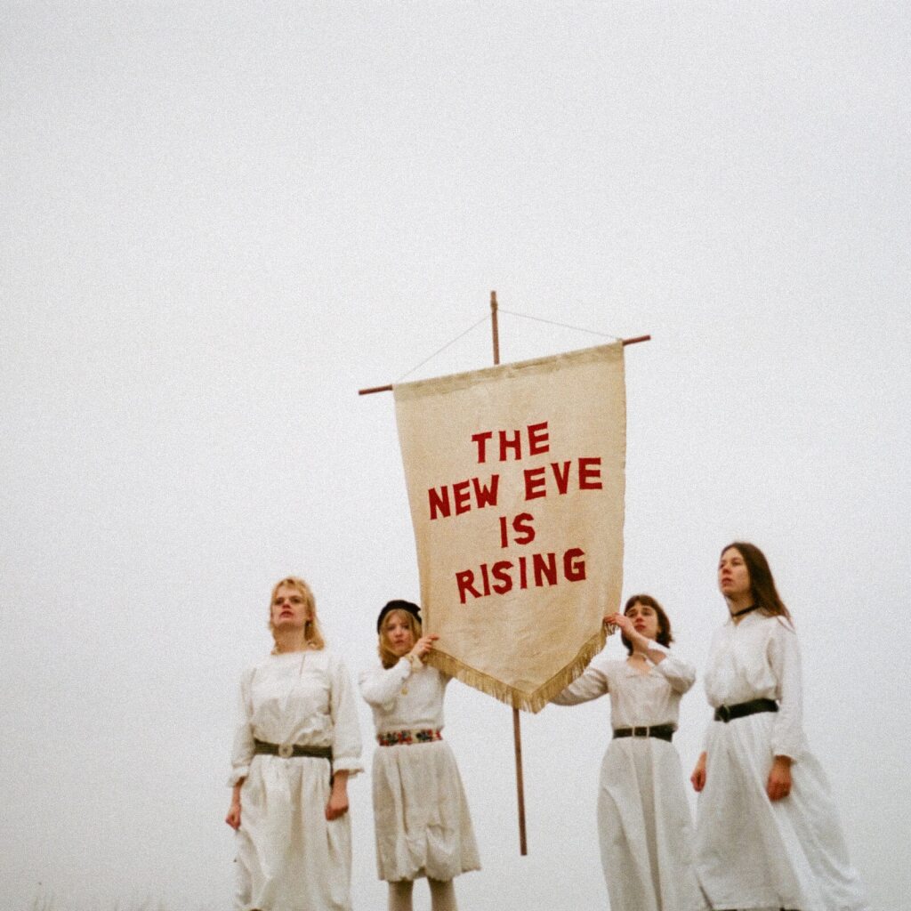 Four women in white dresses hold a banner reading "The New Eve Is Rising.