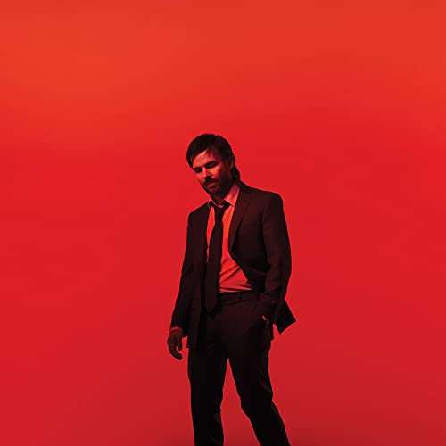 Man in suit against a solid red background, looking downward.