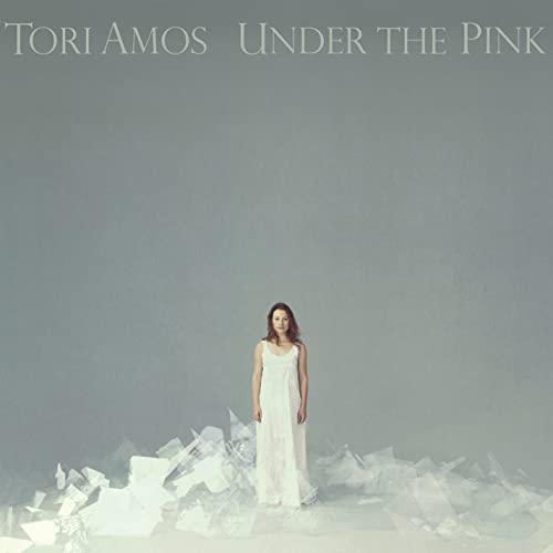 Tori Amos stands in a white dress surrounded by broken glass on grey background.