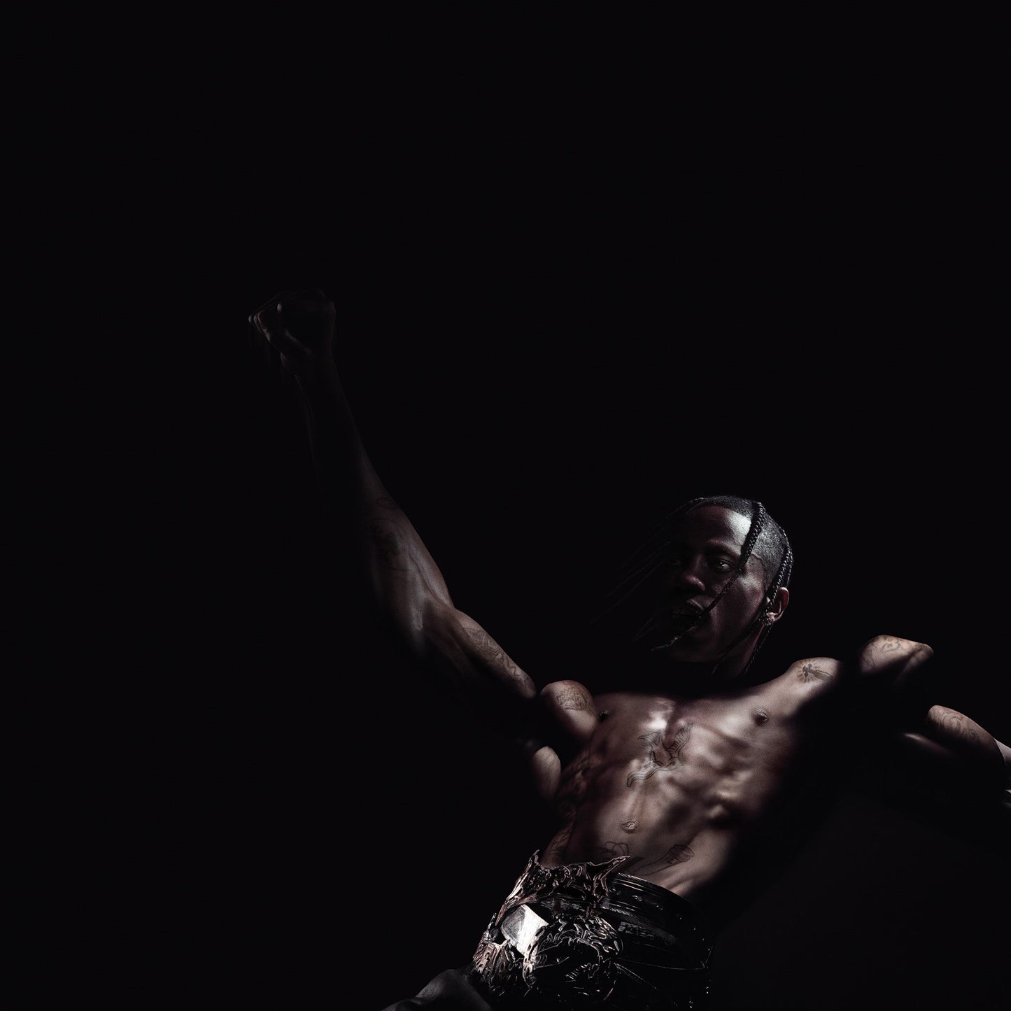 A shirtless man with braided hair poses with one arm raised against black background.