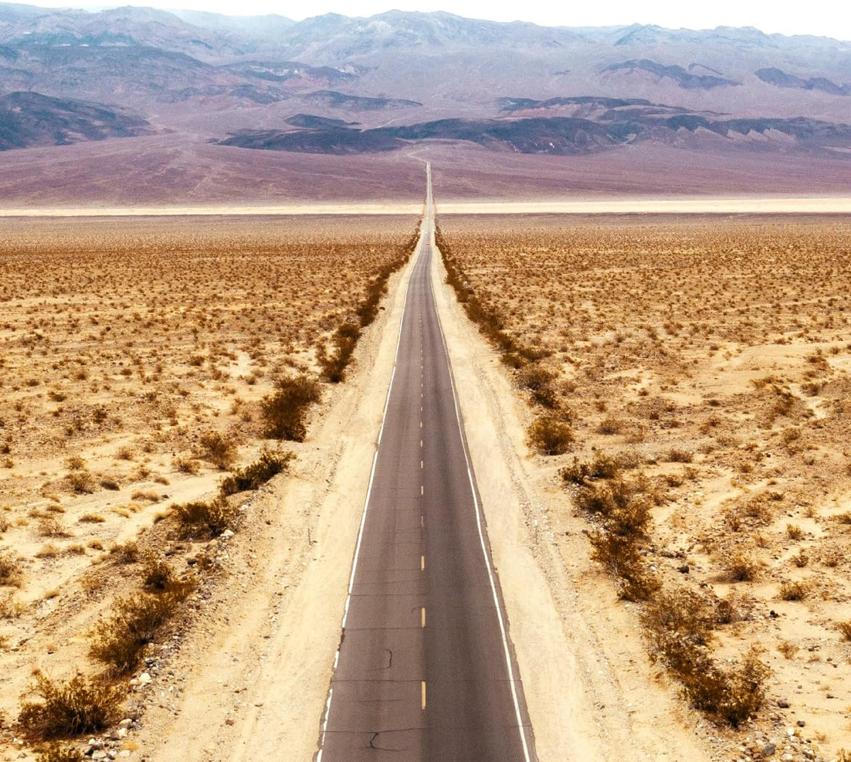Straight desert highway receding towards distant mountains under a pale sky.