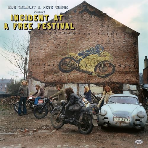 A group of people with motorcycles and a vintage car before a brick wall.