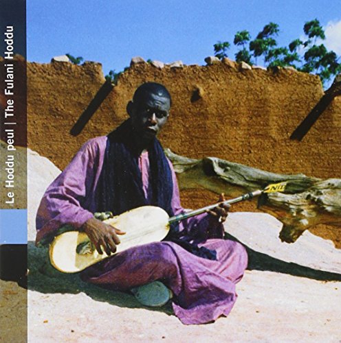 Man playing a stringed instrument, seated against a textured earthen wall.