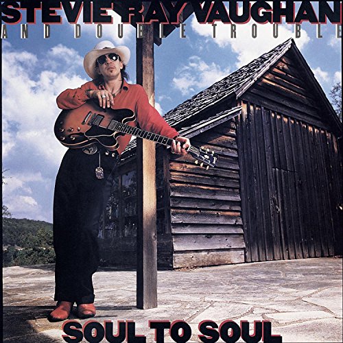 Stevie Ray Vaughan with guitar, leaning against a weathered wooden building.