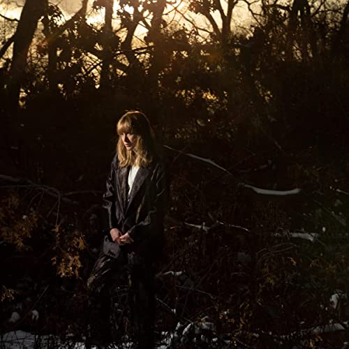 Woman in a black coat stands among bare trees at dusk.