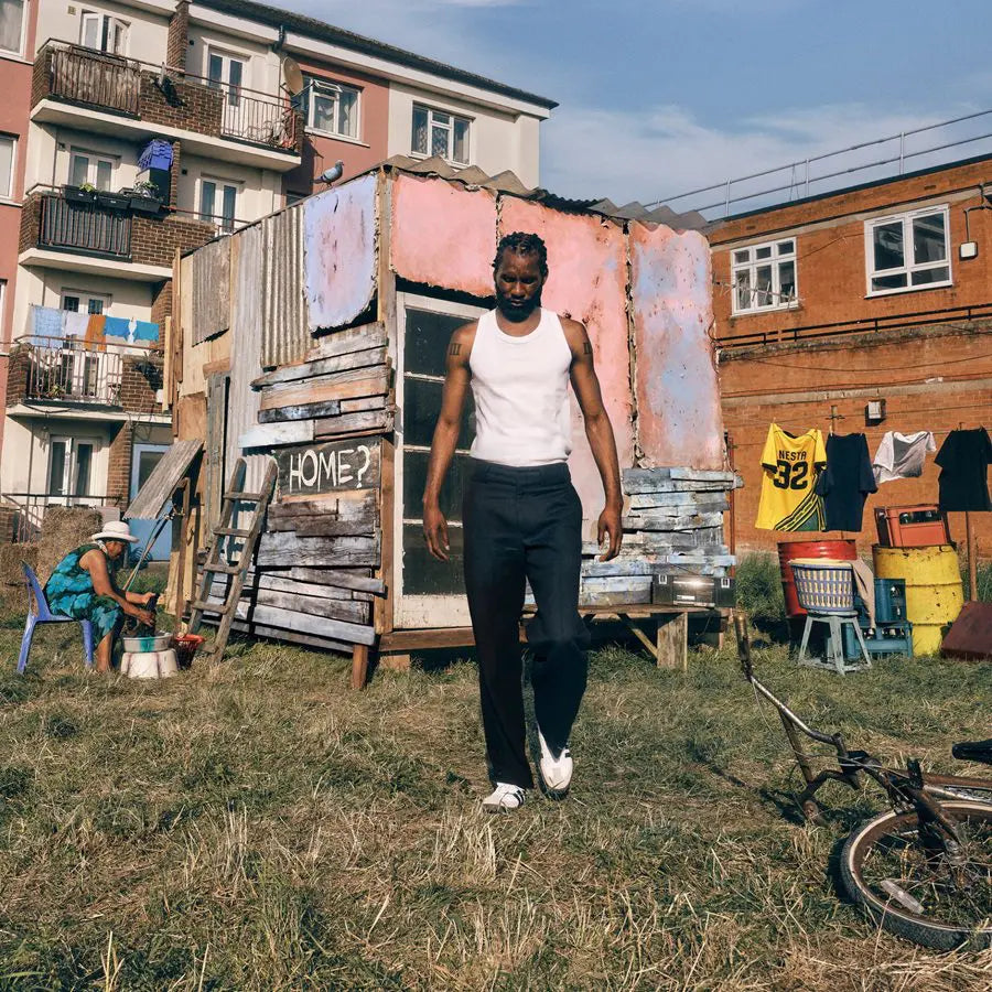 Man standing near a small, weathered building with "HOME?" written on it.