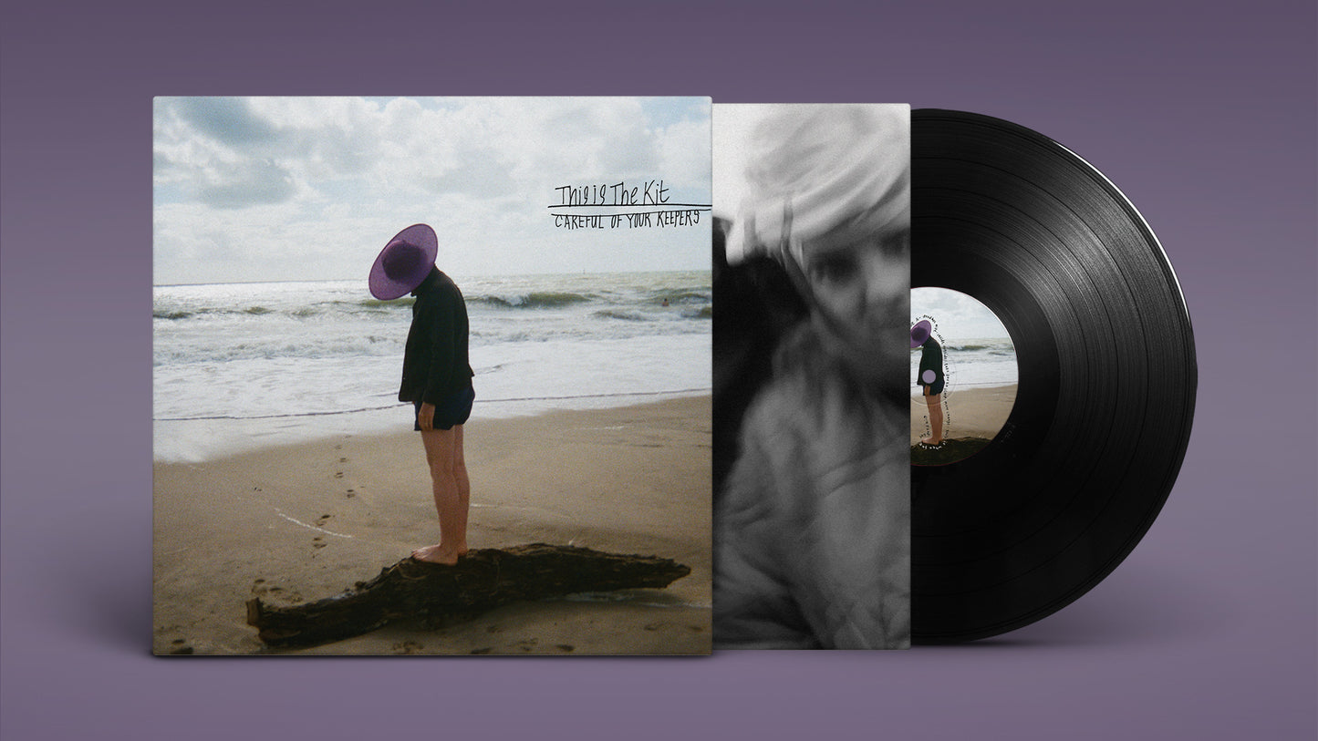 A person wearing a purple hat stands on a log on a sandy beach.