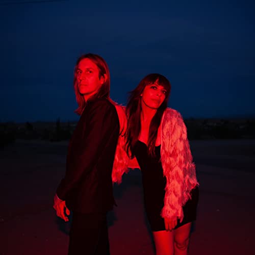 Two people with long hair stand facing away in red and blue light.