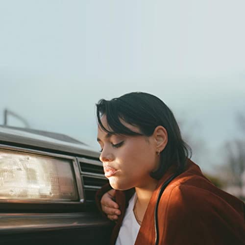 Woman with dark hair leaning against a car, looking downward.