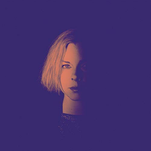 A woman with short blonde hair against a dark purple background.