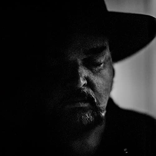 Black and white portrait of Alain Johannes wearing a hat.