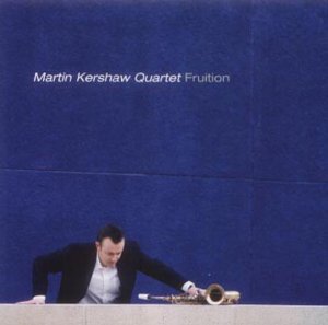 Man in black jacket and white shirt holds saxophone on a table against a blue background.