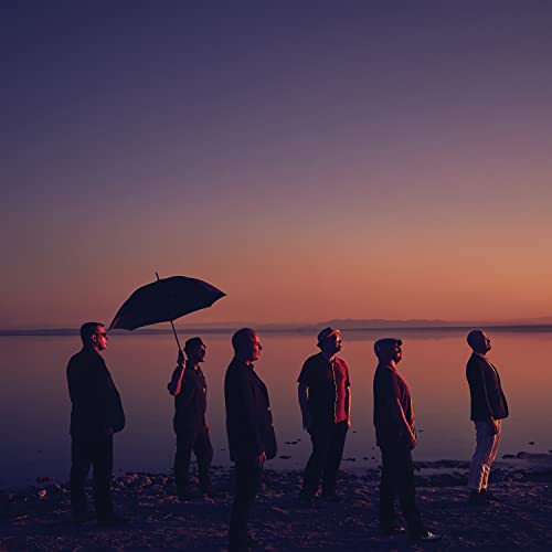 Silhouetted group of six people standing by water at sunset.