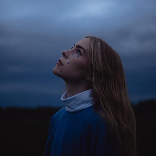 Woman with blue face paint looking upwards against a cloudy sky.