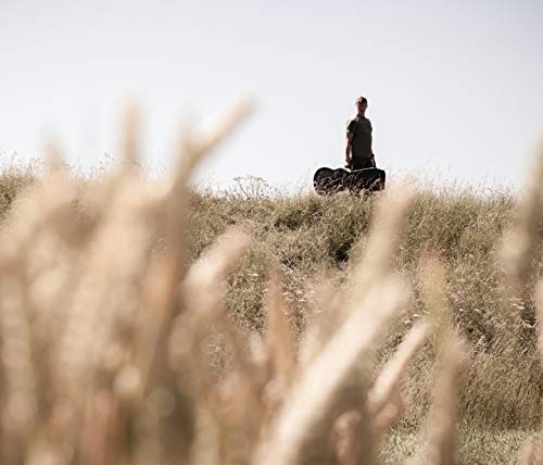 The alt text for the image is: "Man standing on hill with suitcase, surrounded by tall grass."