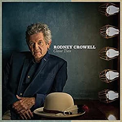 Rodney Crowell sits in a suit next to a beige hat with wire lights.