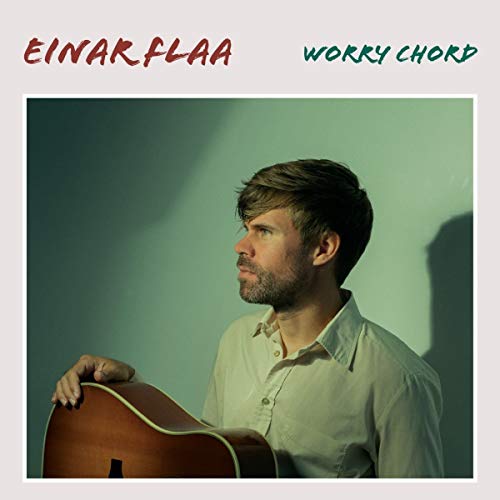 Man holding a guitar, looking left, against a green wall with shadow.