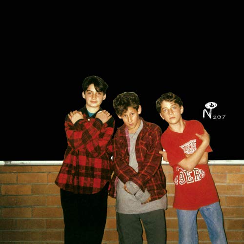 Three young men standing in front of a brick wall against a black background.