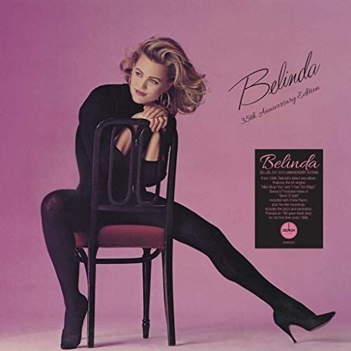 "Blonde woman in black outfit sitting on a chair against a pink background."