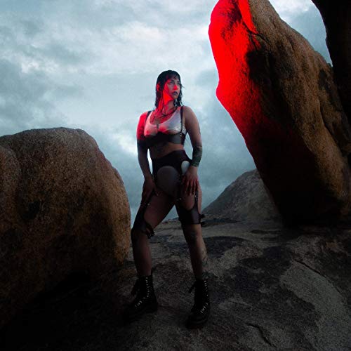 Woman in dark clothing stands among large rocks under a cloudy sky.