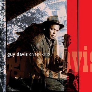 Guy Davis wearing a hat and leather jacket, holding an acoustic guitar.
