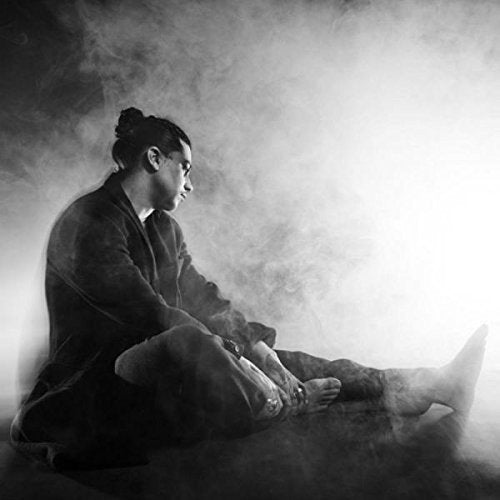 Black and white image of a man sitting, surrounded by smoke, looking to the right.