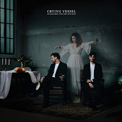 "Album cover: Two men in black sit, woman stands behind, in a dimly lit room."