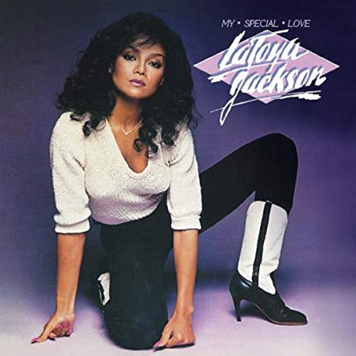 La Toya Jackson kneeling, wearing white top, black pants, and white boots, against purple background.