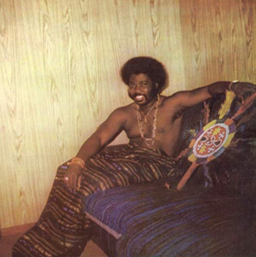 A man sits on a chair, wearing traditional African attire and jewelry, against a wood-paneled wall.