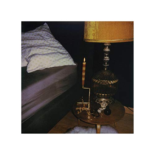 The image depicts a dimly lit bedroom scene. "Bedside table with lamp, lit candle, and skull beside a bed."