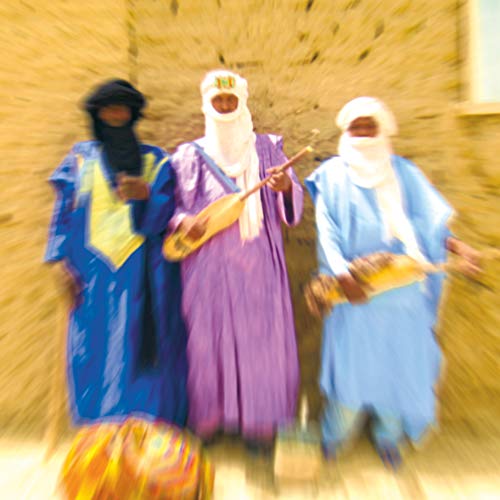 Three men in traditional Tuareg robes and headwraps standing against a textured earthen wall.