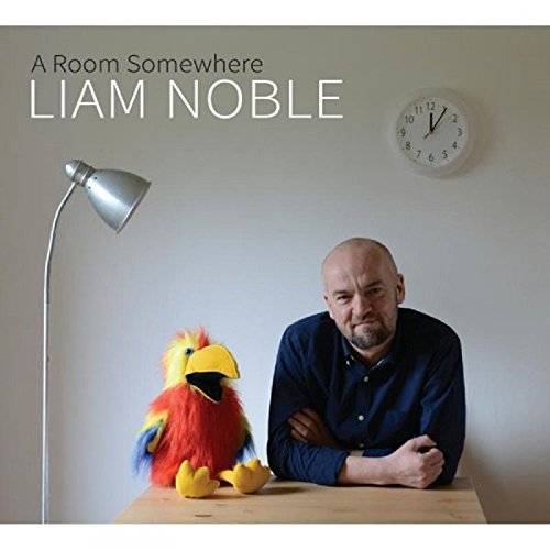 Man with arms crossed sits at table with colorful puppet beside a lamp.