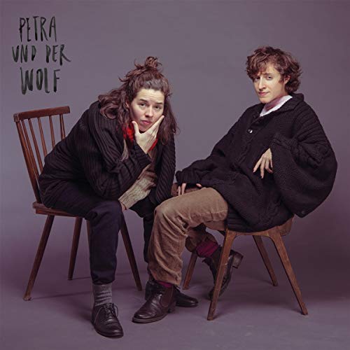 Two people with curly hair sit on chairs, wearing dark coats.