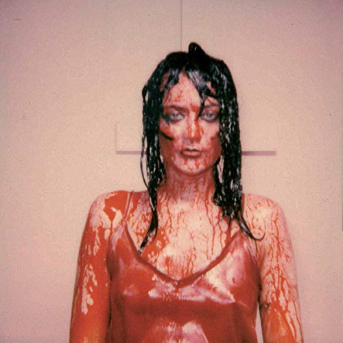 A woman with dark hair and a bloody body stands against a beige wall.