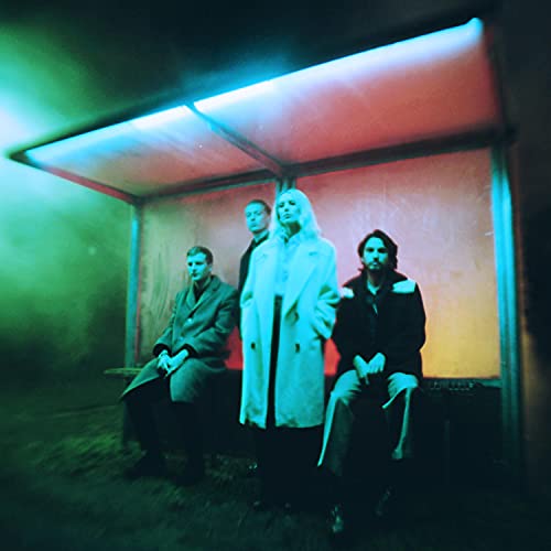 Wolf Alice band members sit at a bus stop under neon lights.