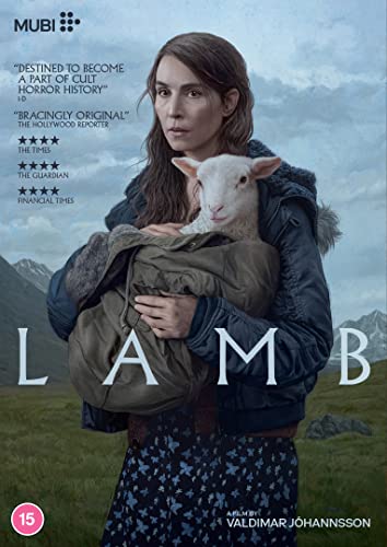 A woman in a dark jacket holds a small white lamb in a rural landscape.