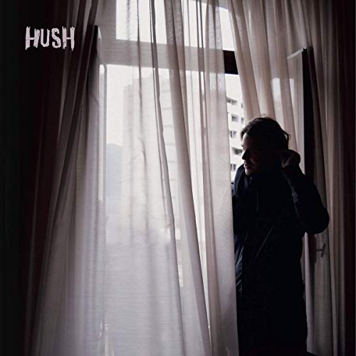 Woman in dark coat stands behind sheer curtains, looking down.