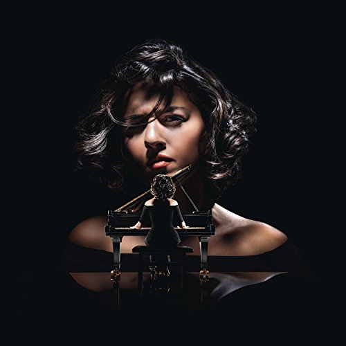 Woman with dark hair playing piano, reflection visible below, against a black background.