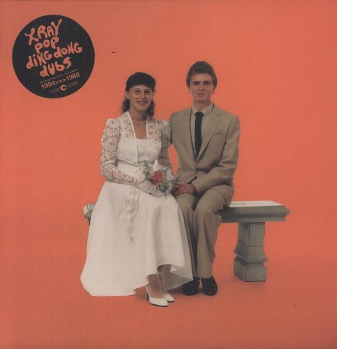 A bride and groom sit on a bench against a solid orange background.
