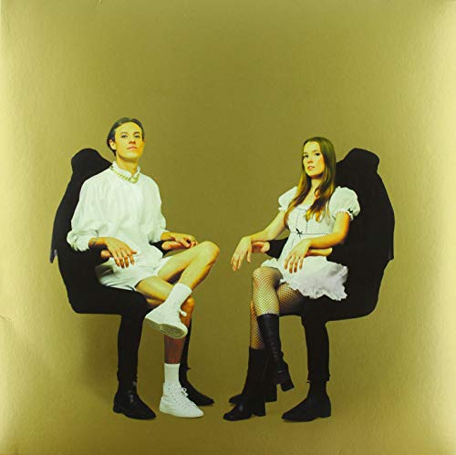 Two people in white clothing sit in black chairs against gold.