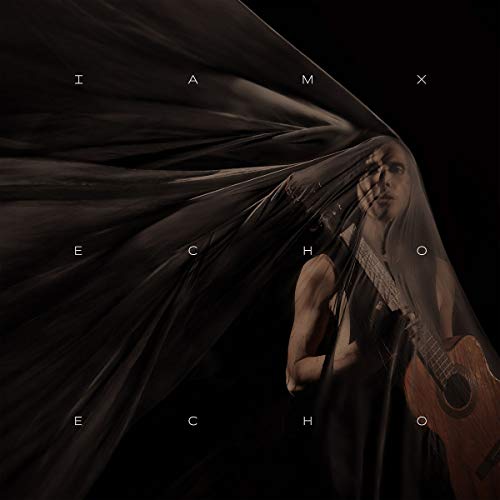 A woman with long hair holds a guitar, partially obscured by flowing black fabric.