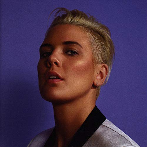 The image shows a woman with short blonde hair against a purple background. Alt text: Woman with short blonde hair and grey shirt against purple background.