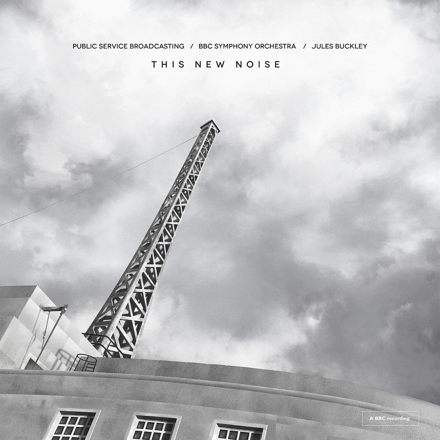 "Black and white image of a tall metal tower against a cloudy sky and building."