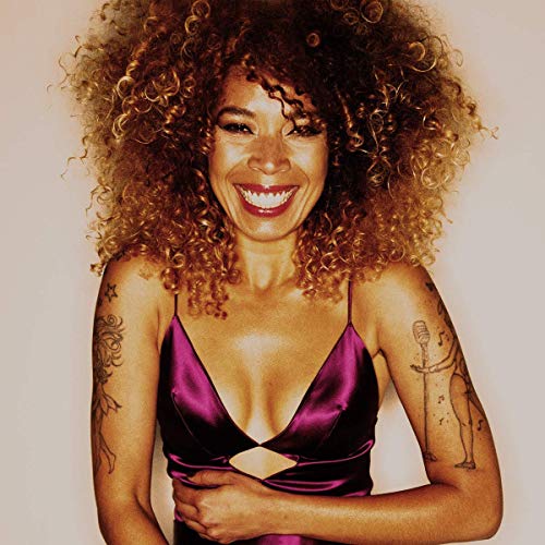 Flavia Coelho with curly hair and tattoos wearing a purple satin top smiling.