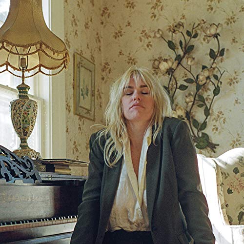 "Blonde woman in a dark blazer and white shirt stands in a floral bedroom."
