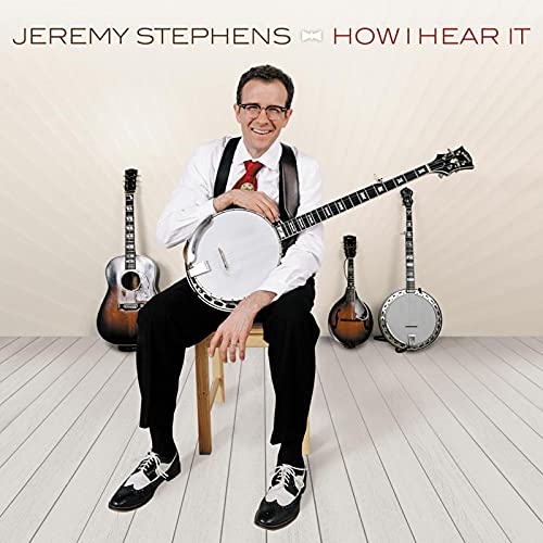 Man sitting on stool holding banjo, surrounded by guitars and banjos, on a white floor.