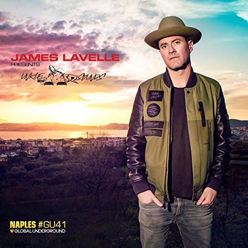 Man in green jacket and hat stands before a cityscape at sunset.