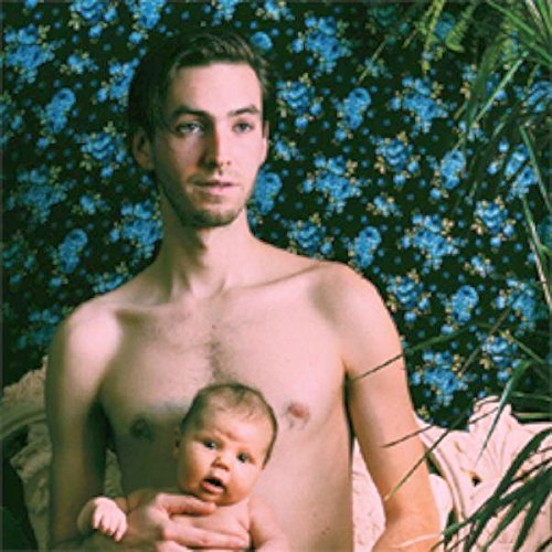 A man holding a baby, both shirtless, surrounded by foliage and blue flowers.