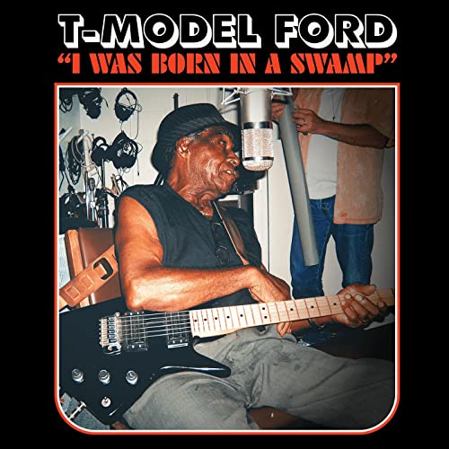 T-Model Ford seated, playing a black electric guitar in a recording studio.